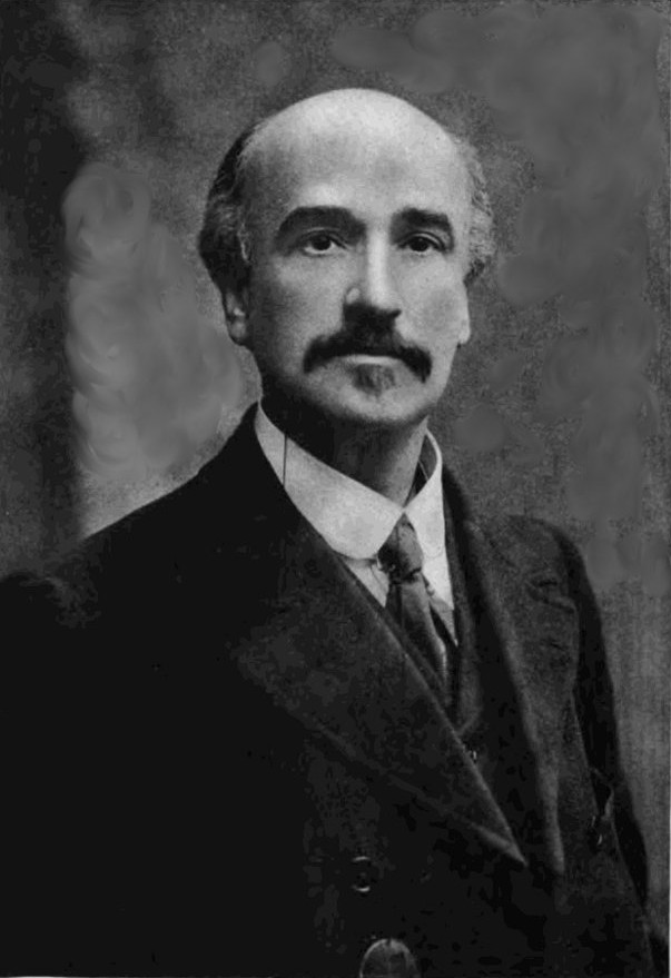 A black-and-white portrait of a man with a prominent mustache and a receding hairline, dressed in a formal suit with a tie. He has a serious expression and is looking directly at the camera, set against a softly blurred background.