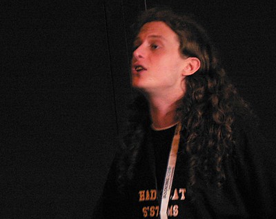 The image shows a person with long, curly hair, speaking passionately while wearing a black shirt with text on it. The background appears dark, suggesting a stage or presentation setting.